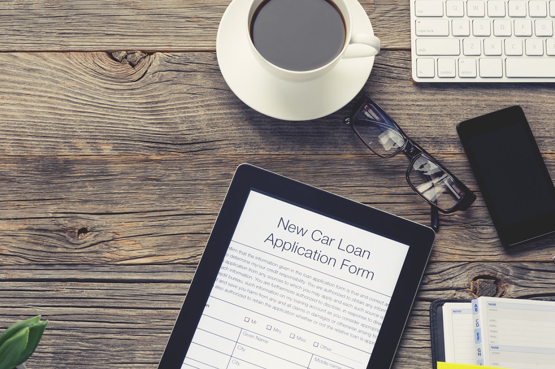 Online car loan application form. The document is displayed on a digital tablet. There is also a mobile phone, computer keyboard, coffee, Filofax organizer notepad and glasses on the table. The table is made of wood and is quite old. Copy space on left.